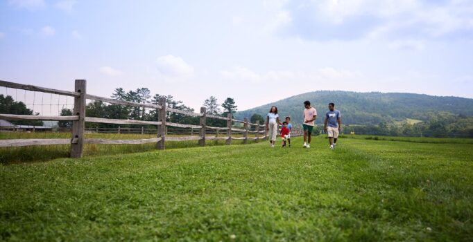 A group of adults and children walk together along a grassy path next to a rustic wooden fence. The wide-open field is bordered by lush green hills and a bright, clear sky in the background.