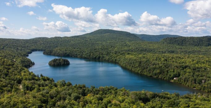 An aerial, wide-angle view shows a deep blue lake nestled among rolling, densely forested green mountains under a clear sky. A small, tree-covered island sits near the center of the lake.