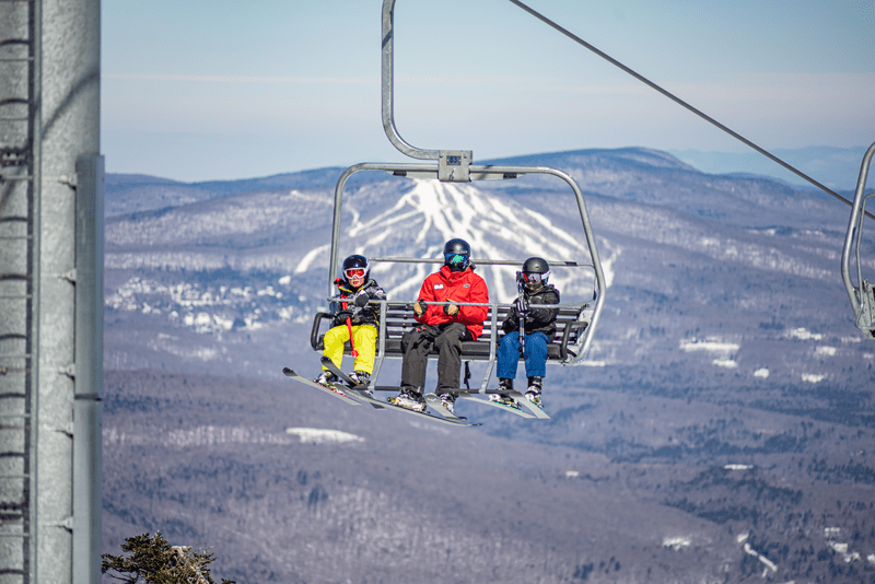 Three people on a chairlift with mountains visible behind on a sunny day.