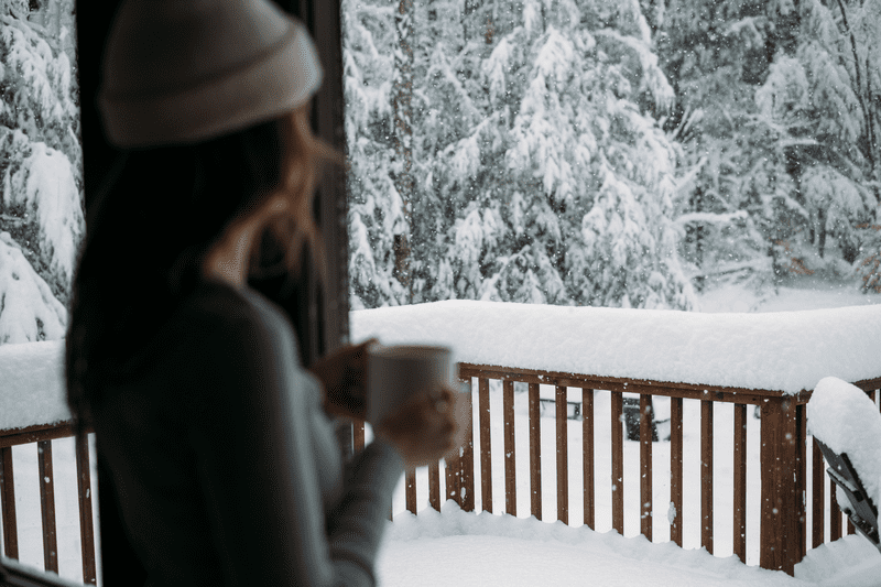 A person looks out onto a snowy deck and forest while holding a mug in their hands.