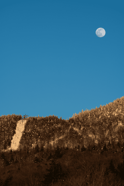 The view from below of a ski run on a mountain. The sky is a deep blue and the moon is visible.