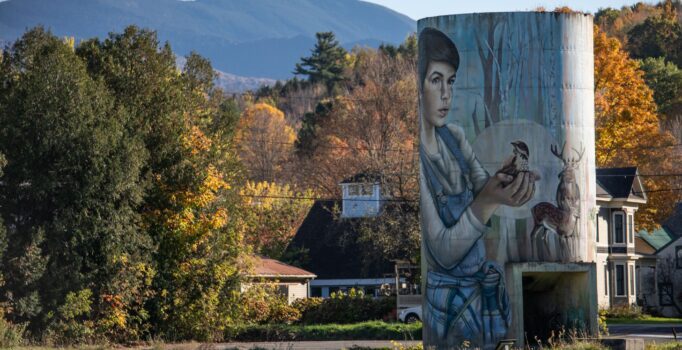 A concrete silo mural that depicts a young child with wide eyes cradling a tiny bird in their hands. The silo is framed by the brilliant yellows of autumn foliage, with soft blue mountains stretching across the horizon.