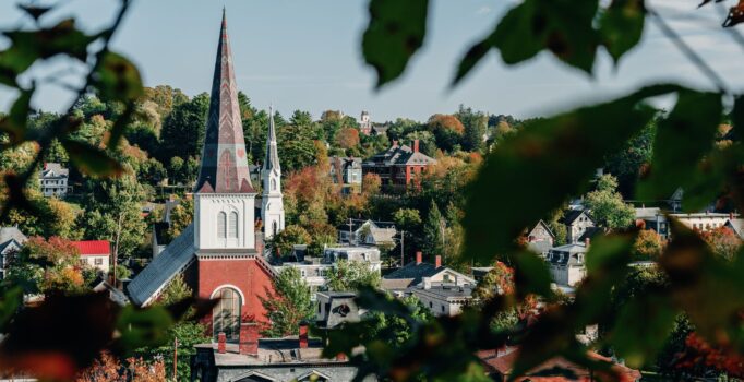 Tall, brick buildings and church steeples rise above the tree line on a sunny day.