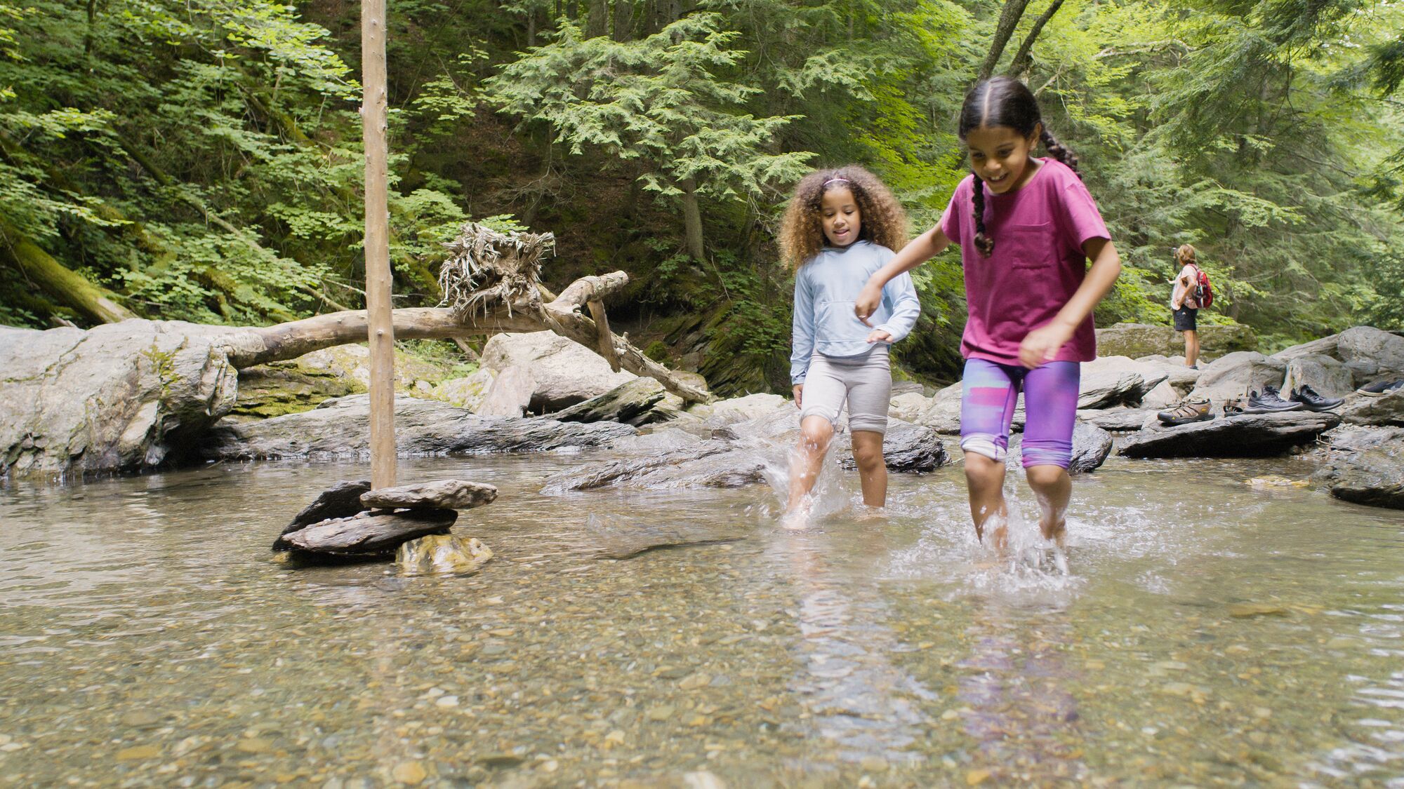 Two children run through a shallow stream in the woods on a warm day.