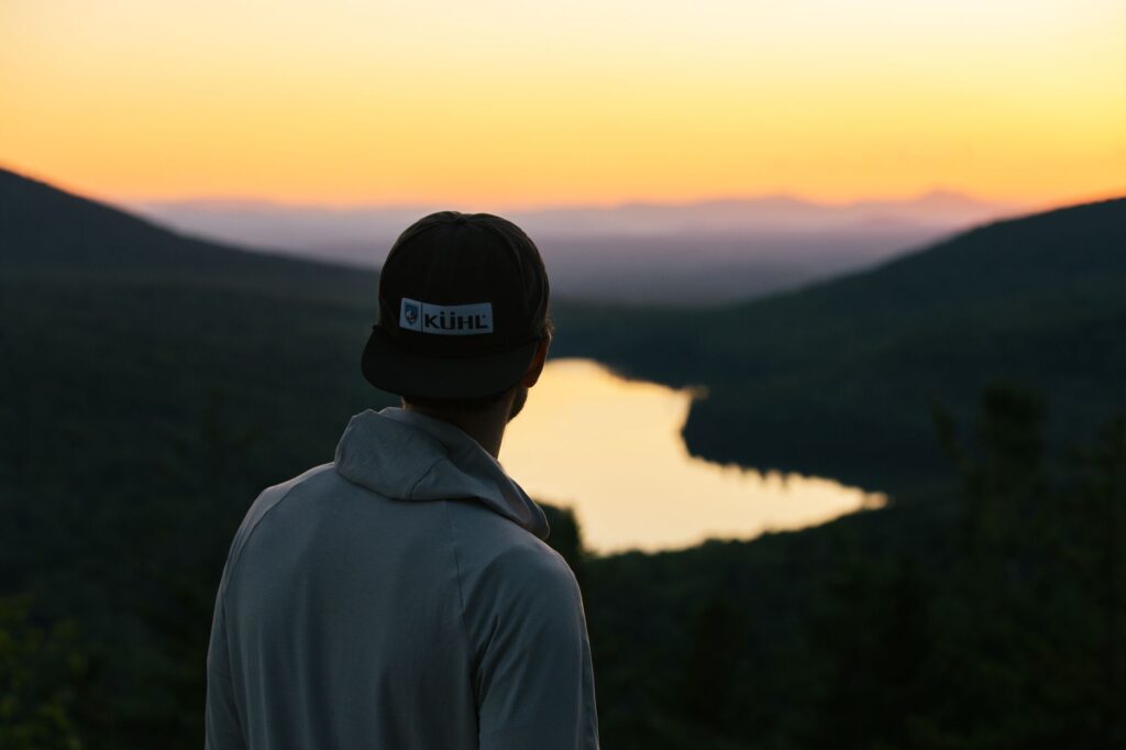 Seen from behind, a person looks at the view of a lake below them with the sunset in the distance.