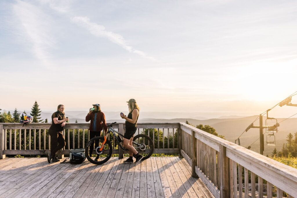 Three people stand and talk on a deck at the top of a mountain while the sun is setting.
