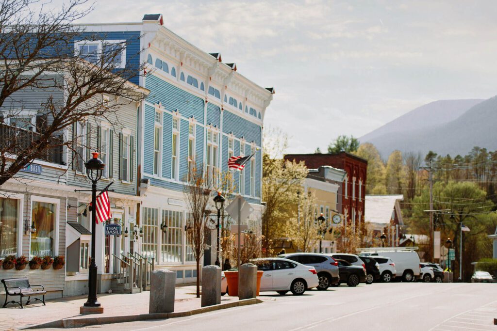 Street view of a downtown area with shops lining the side of a road and mountains in the background.