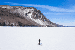 A cross-country skier skis across a frozen lake with a rocky ledge in the background.