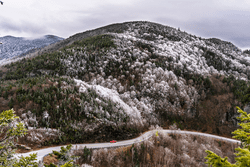 Seen from far above, a red car drives on a road that’s wrapped around a mountain with snow dusting the treetops.