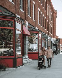Two people pushing a baby in a stroller walk through a historic downtown in the summer.