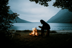 A person kneels by a campfire next to a lake on a dark, cloudy day.