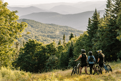 Three people with mountain bikes pause at a clearing in the woods to take in an open view of mountains.