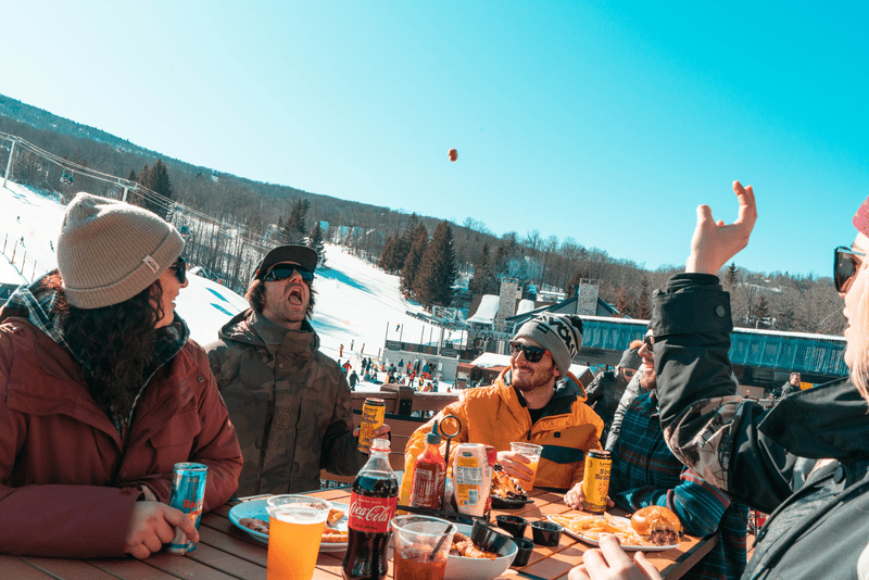 A group of five people eating and drinking at an outdoor table at a ski area.