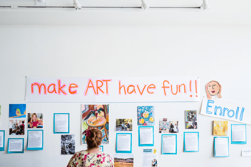 A person looks at a group of illustrations on a wall and a sign that reads Make Art Have Fun!!