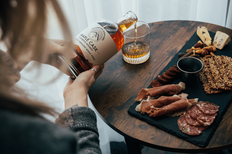 A person pours from a bottle of bourbon whiskey into a cocktail glass with charcuterie alongside.