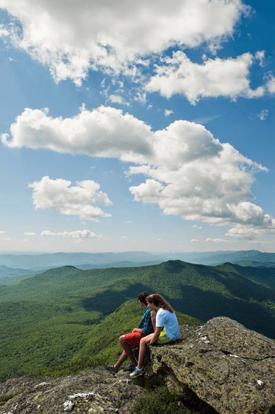 Two people sit on a rock at the top of a mountain and look at the view of other mountain peaks.