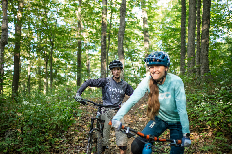 Two people riding their bikes through a forest smile for the camera.