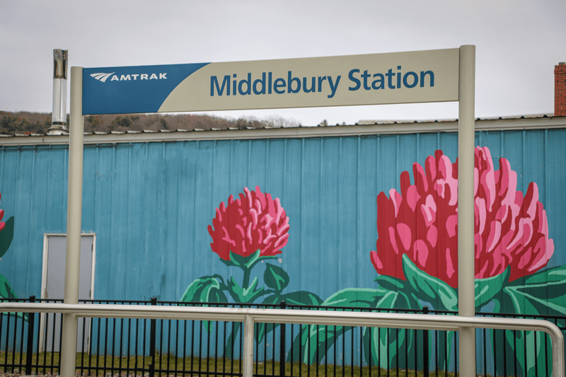 A mural of flowers is painted on a building behind a sign that reads Amtrak Middlebury Station.