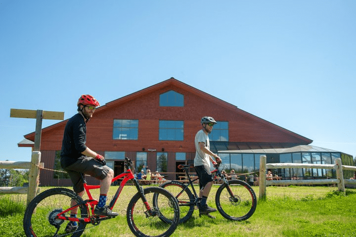 Two people stand with their bikes next to a large barn on a sunny day.
