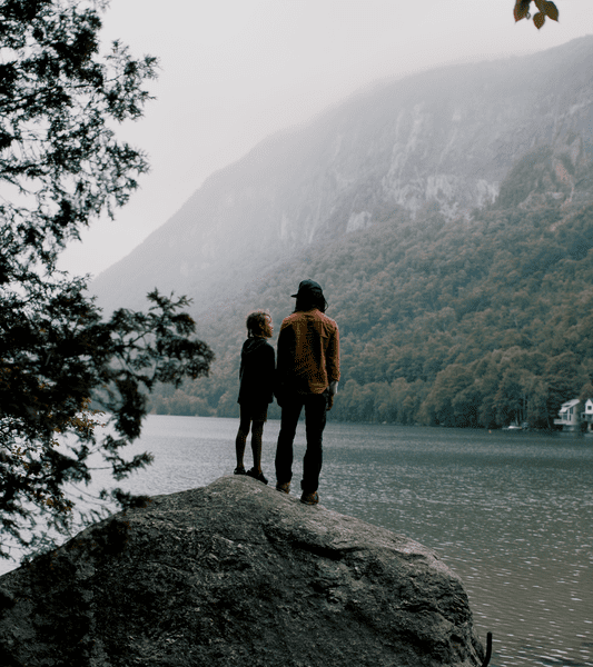 Seen from behind, two people stand on a rock facing a lake on a foggy day.