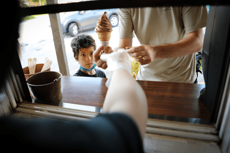 Soft serve ice cream is served on a cone from an ice cream truck to an adult and child.