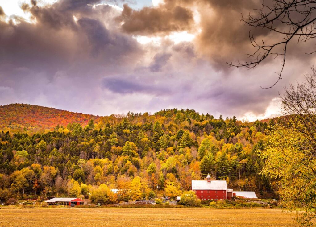 A red barn in the fall with red, orange, and yellow leaves visible on trees on a mountain behind it.