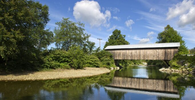 On a sunny day, a wooden covered bridge is reflected on the river running below it.