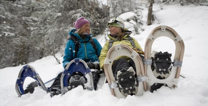 Two people sit in the snow wearing their snowshoes.