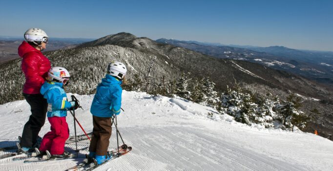 A family with children pause at the top of a snowy ski run with mountains surrounding.