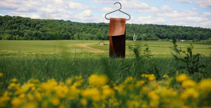 A metal piece of art with a hangar atop a column, sits outdoors in an open, green field on a sunny day.