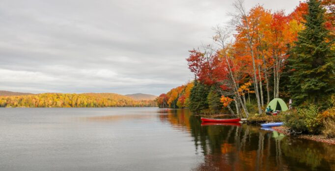 A canoe is tied up next to a camp site on the shore of a calm body of water, surrounded by fall foliage.