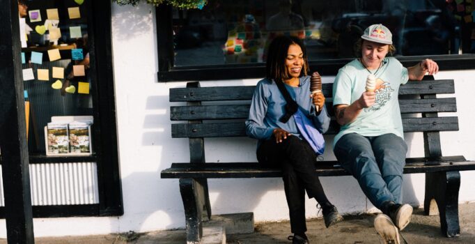 Two people sit on a bench, each holding a cone with soft ice cream.