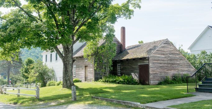 An old wooden home is surrounded by a grassy lawn and a wooden fence.