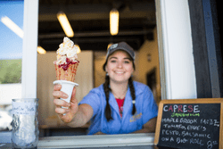 A person with dark hair in two braids working behind a counter holds out an ice cream cone to the camera. It has various colorful toppings on it.