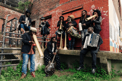 A nine-piece band photographed outside in the summer.