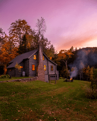 A large cabin in the woods seen from outside. A fire crackles in an outdoor fire pit and the windows emit warm light.