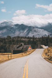 A road without traffic leading past a barn with a snowy mountain in the background.