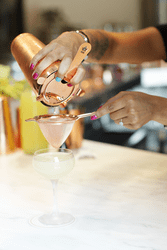 Hands with brightly colored nails pour a drink from a shaker into a glass.