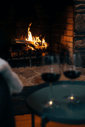 Two wine glasses sit in front of a fireplace with a fire.