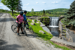 Two people stand with their bikes on a dirt path to look at a waterfall.