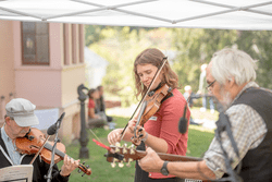 Three people play instruments under a tent outdoors on a warm day.