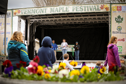 People look at an outdoor stage in the spring with a sign that reads Vermont Maple Festival.