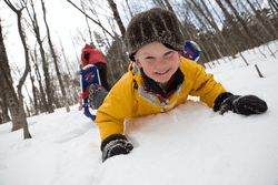 A child, wearing snowshoes, smiles at the camera while laying on their chest in the snow.