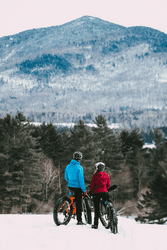 Two people facing away from the cameras standing on bikes with fat tires admiring a mountain view in the winter.
