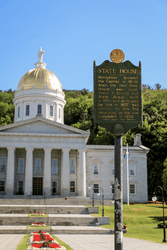In front of a historic building with a golden dome, a green roadside marker reads State House.