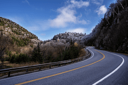 A road winds through a mountain range with snow dusting the tops of the trees.