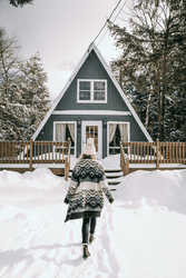 Seen from behind, a person walks toward a snow-covered a-frame house in the winter.