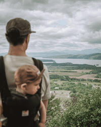 A person carries a young child in their backpack while looking at the view from a mountain.
