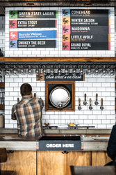 A person facing away from the camera pours a beer from a tap on a wall with white subway tile covering it. Above them is a list of beers on tap.