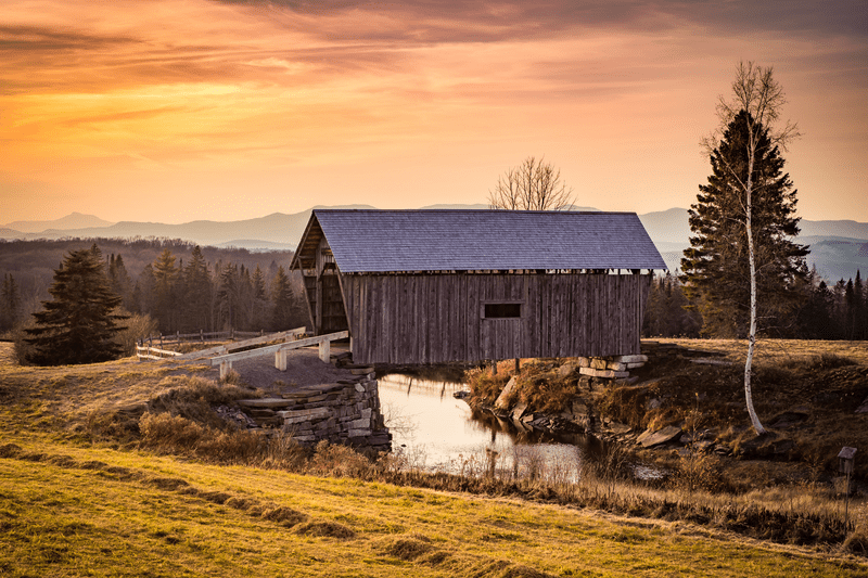 A covered bridge in the countryside as the sun sets with mountains visible behind.